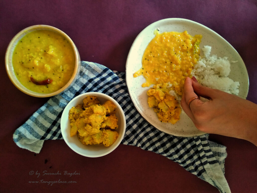 served plate of tok dal rice and aloo poshto with bowlfuls of tok dal and aloo poshto on checkered cloth and maroon background