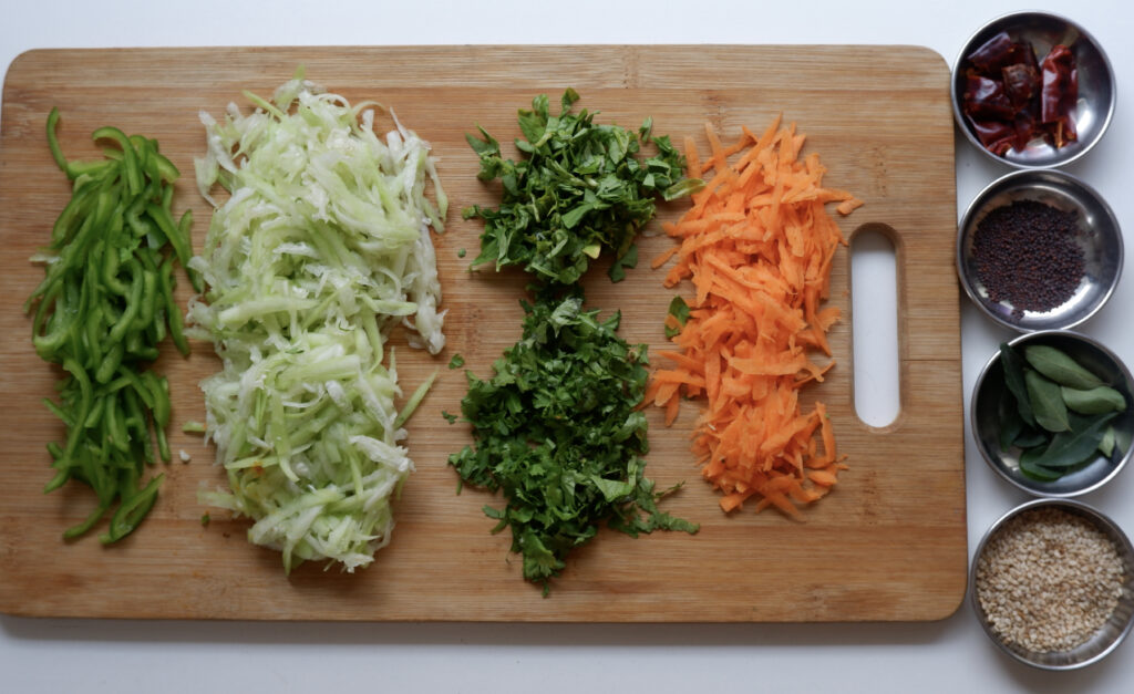 Chopping board with grated capsicum, bottle gourd, coriander, curry leaves and carrot along with small plates of mustard seeds, curry leaves, sesame seeds and broken red chillies