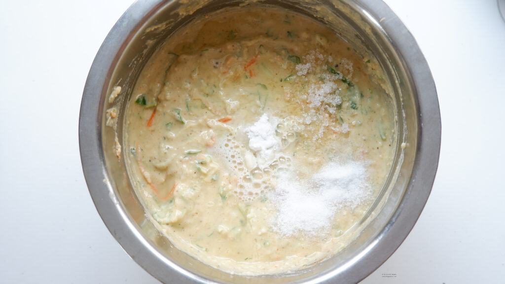 A steel container containing grated vegetables added Handvo batter on a white back ground